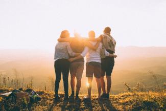 four person hands wrap around shoulders while looking at sunset by Helena Lopes courtesy of Unsplash.